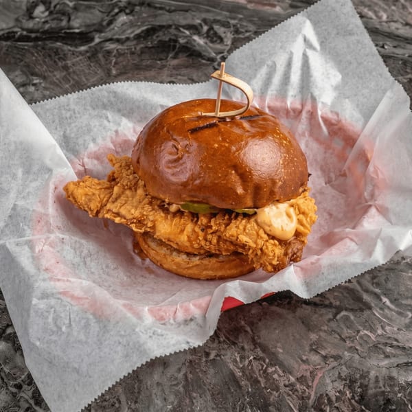 Crispy fried chicken sandwich with sauce and pickles on a brioche bun, served in a paper-lined basket.