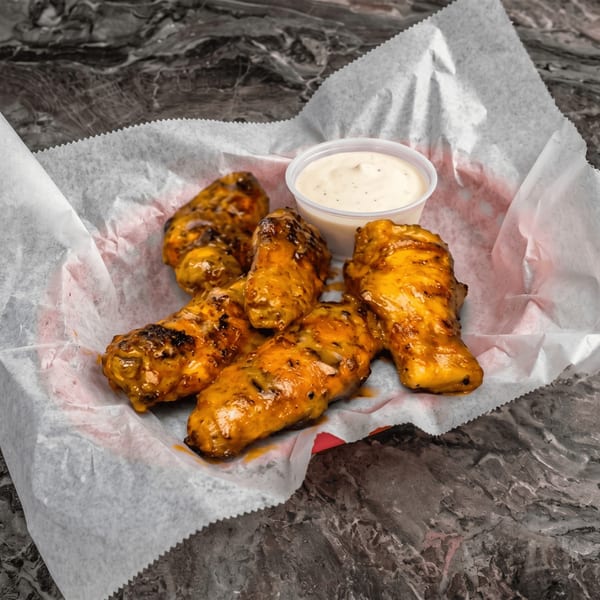 Five golden-brown chicken wings with sauce in a paper-lined basket, with a side of ranch.