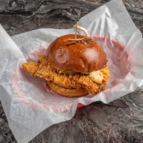 Crispy fried chicken sandwich with pickles and sauce on a brioche bun, served in a paper-lined basket.